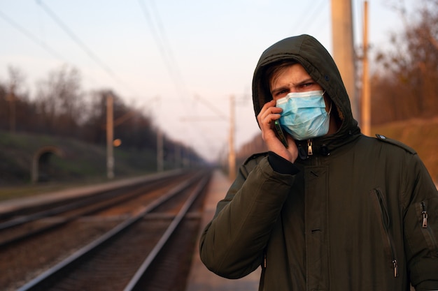 Man met een telefoon in zijn handen te wachten op een trein in een medisch masker vanwege de verspreiding van coronavirus, virusuitbraak, luchtverontreiniging.