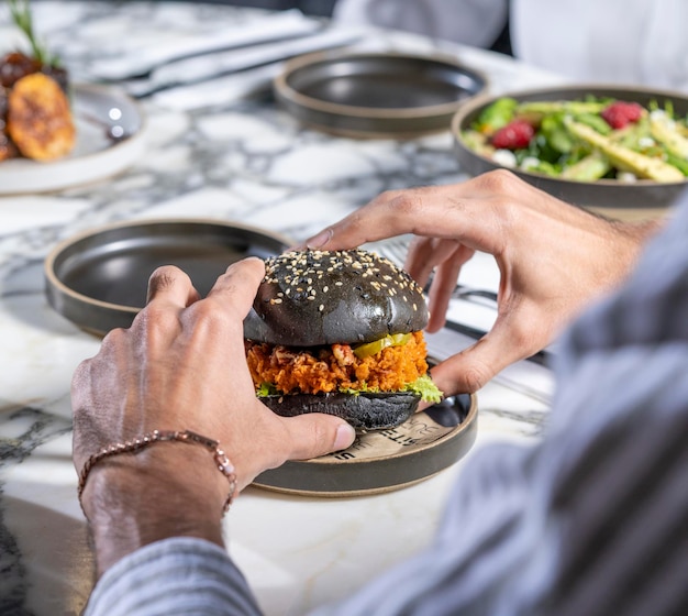 Man met een zwarte broodje kipburger met een witmarmeren tafel op de achtergrond.