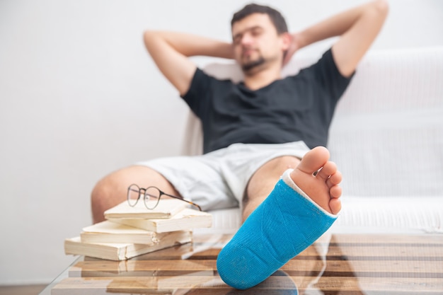 Man met gebroken been in blauwe spalk voor de behandeling van verwondingen door enkelverstuiking die boeken lezen bij de revalidatie thuis.