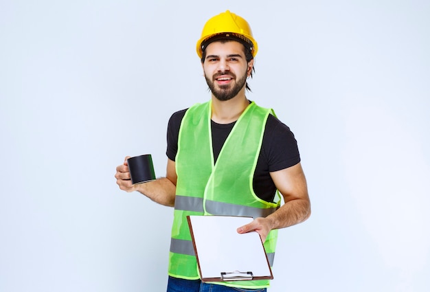 Man met gele helm met projectmap en een kopje koffie.