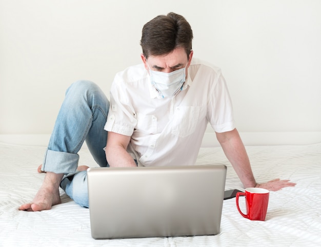 Man met gezichtsmasker met behulp van laptop op bed. Werk vanuit huis tijdens quarantaine en zelfisolatie. Coronavirus concept