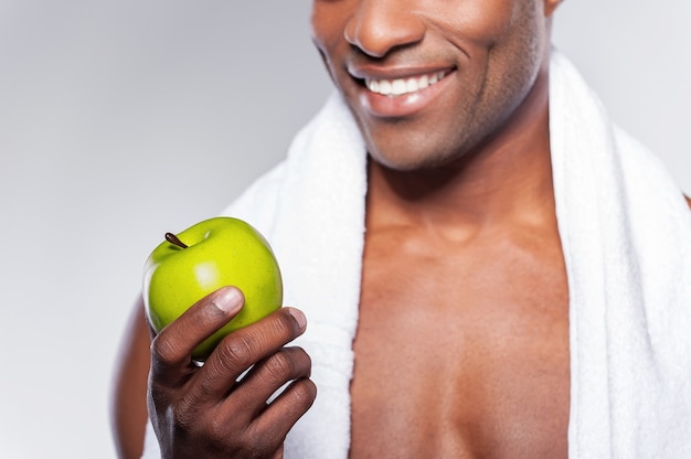 Man met groene appel. Bijgesneden afbeelding van jonge gespierde Afrikaanse man met handdoek op schouder die een appel overgeeft en naar de camera glimlacht terwijl hij tegen een grijze achtergrond staat