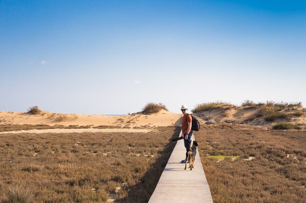 Man met hond die op het houten pad op het strand loopt en in de verte van de oceaan kijkt.