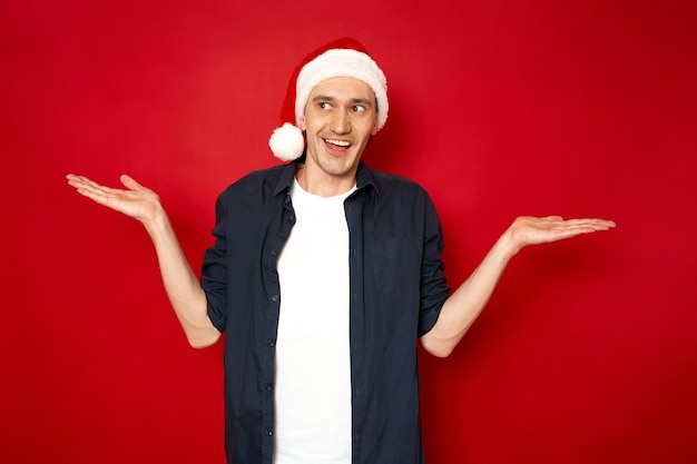 man met kerstmuts verbaasde uitdrukking spreidt handen glimlacht absurd geïsoleerde rode achtergrond
