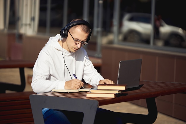 Man met koptelefoon werkt op een laptop en schrijven in een notitieblok zittend op straat aan tafel