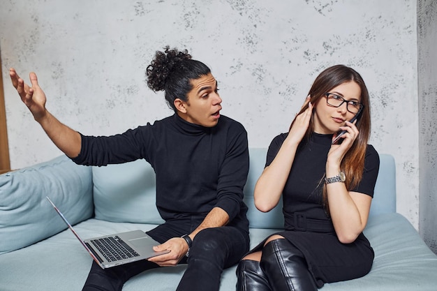 Man met krullend haar en vrouw die in zwarte kleren met laptop binnenshuis zit. Kerel is boos.