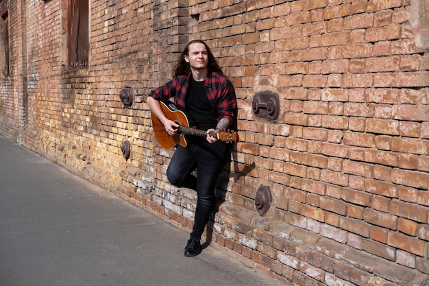 Man met lang haar die gitaar speelt leunt tegen een bakstenen muur op straat