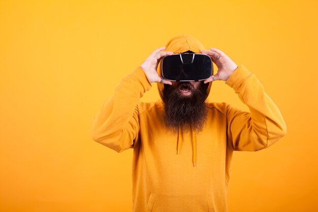 Man met lange baard met een virtual reality-headset die geschokt kijkt over gele achtergrond. Gele hoodie. Moderne technologie. Knappe man.