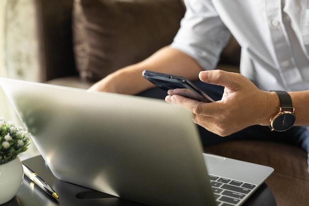 Man met mobiele telefoon en laptop thuis zittend op de bank in de woonkamer thuis