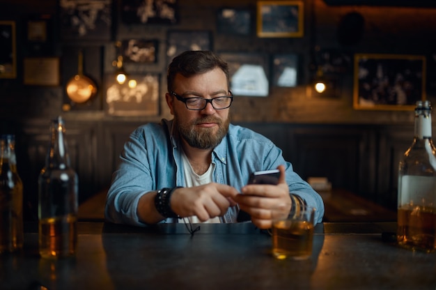 Man met mobiele telefoon zit aan de balie in de bar. Een mannelijke persoon rust in de kroeg, menselijke emoties, vrijetijdsactiviteiten, nachtleven