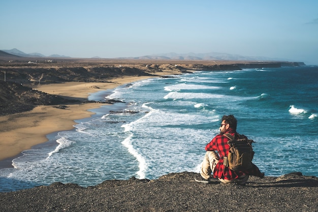 Man met rugzak gaat op de klif zitten en geniet van het lege wilde strand met grote oceaangolven