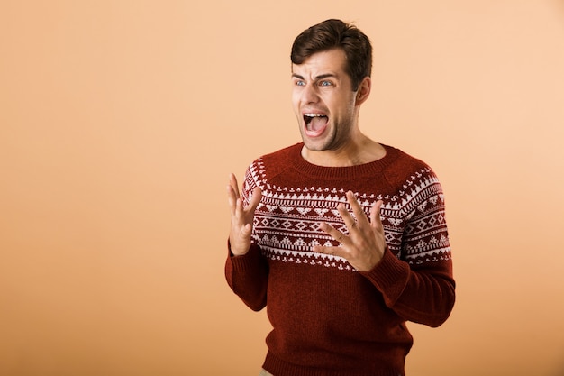 Man met stoppels die gebreide trui draagt die en verontwaardiging schreeuwt, geïsoleerd over beige muur