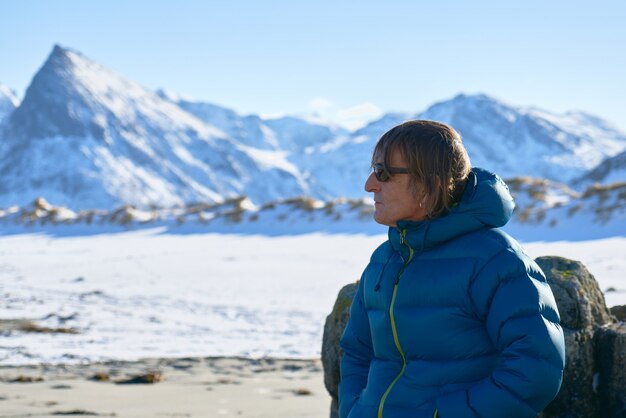 Man met zonnebril kijkt opzij. Met sneeuw bedekte bergen en strand op de achtergrond.