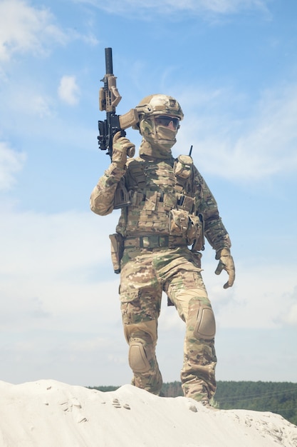 Man in military camouflage uniform and mask, equipped tactical ammunition, standing on sand dune with service rifle replica in hands, cloudy sky on background. Airsoft player taking part in war games