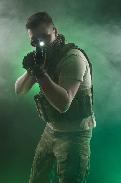 The man in military special clothes posing with a gun in his hands on a dark background in the haze