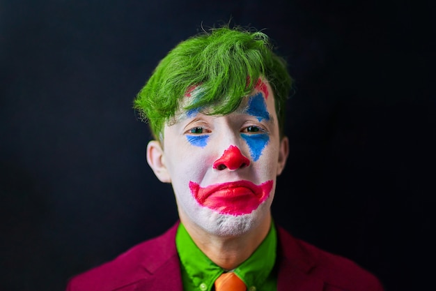 Man in mime makeup cosplay with green hair and a red suit an orange tie and a green shirt clown sad