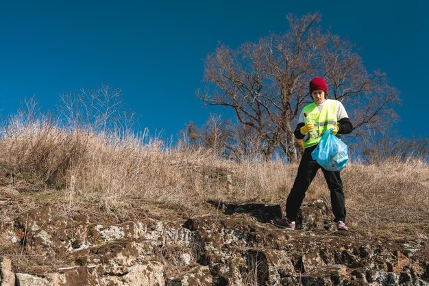 Man nature activist in yellow vest puts glass bottle in the\
blue trash bag outdoors