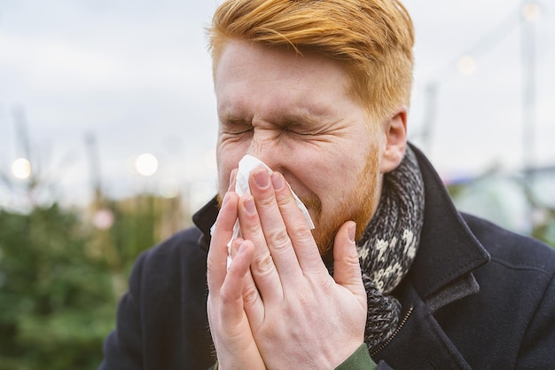 man niest en snuit zijn neus in een zakdoek in de winter met allergie of een infectie een virus. Portret van roodharige man buiten.