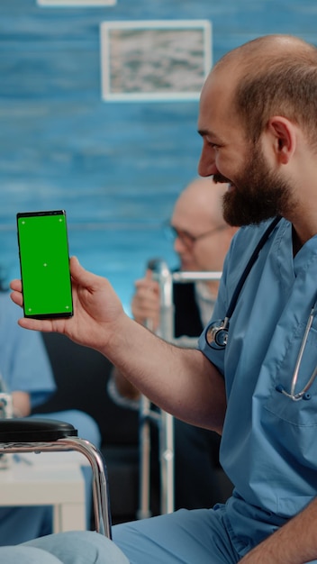看護施設でスマートフォンの緑色の画面を保持している男性看護師。車椅子に座っている老婆とモックアップテンプレートと孤立した背景のクロマキーを見ている医療助手