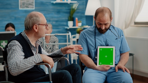 Man nurse vertically holding green screen on tablet while consulting patient with disability. Retired person in wheelchair looking at chroma key with isolated mockup template on device.