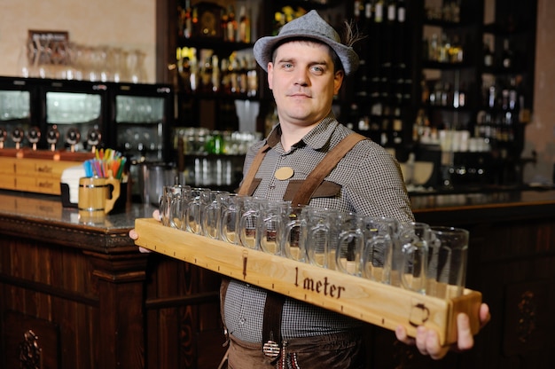 A man at the Oktoberfest festival in traditional Bavarian clothes and a hat holds a meter of beer and smiles