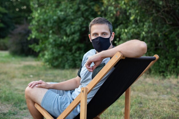 Man ontspannen op de ligstoel in beschermend medisch masker op zijn gezicht zonnebaden buiten groen gazon in park