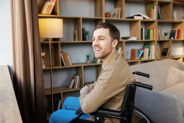 Man op rolstoel in de kamer kijkt door het raam en glimlacht Zijaanzicht van een jonge gehandicapte patiënt in een rolstoel die uit het raam kijkt