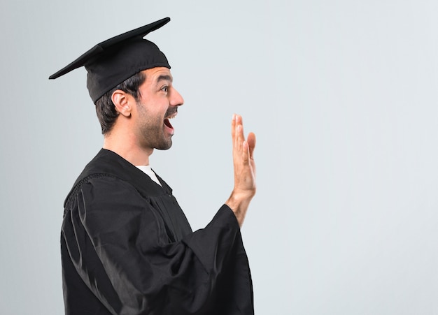 Man op zijn afstuderen dag Universiteit groeten met de hand met gelukkige uitdrukking op een grijze achtergrond