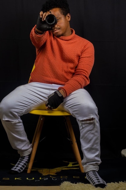 A man in an orange sweater sits on a yellow chair with a black background.