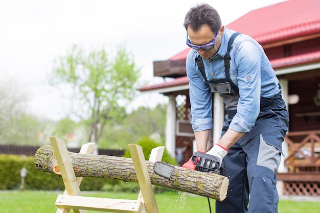 Man in overalls saws a tree on sawhorses in courtyard with a chainsaw