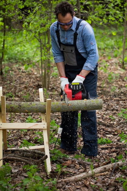 Man in overalls saws wood with chain saw using sawhorse in woodland