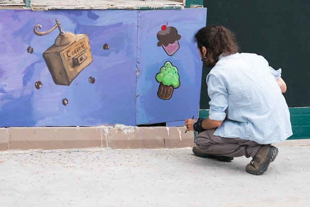 Man painter with beard draws coffee and cakes on the lateral side of the stage