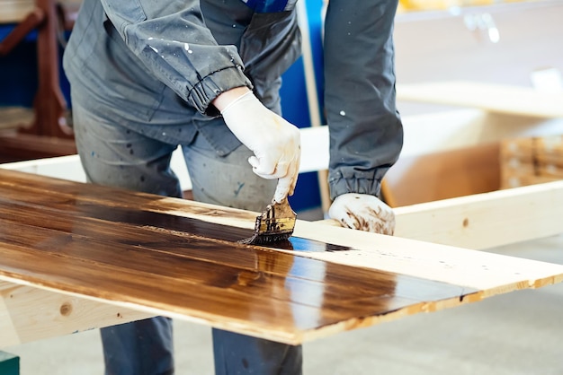 Man paints wooden boards with paint brush Carpenter cabinetmaker varnishes wooden surface Authentic workflow