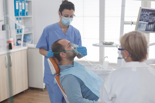 Man patient sitting on dental chair during stomatology\
examination while dentist assistant putting oxygen mask before\
teeth surgery in stomatological office room. medical team wearing\
face mask