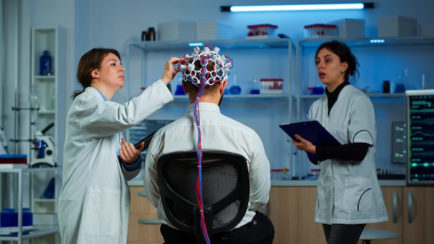 Photo man patient wearing performant eeg headset scanning brain electrical activity in neurological research laboratory while medical researcher controlling it, examining nervous system, nurse taking notes