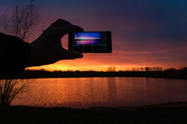 写真 スマートフォンで湖の上の日没を撮影する男性