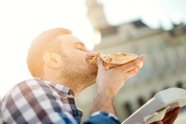 Man pizzasnack buitenshuis eten. Knappe jonge man die een stuk pizza buiten op straat eet.