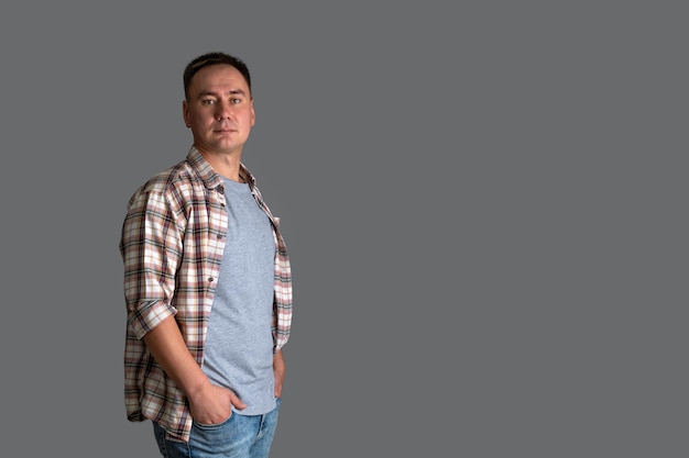 A man in a plaid shirt and a gray T-shirt stands on a gray background with his hands in his pockets, copyspace