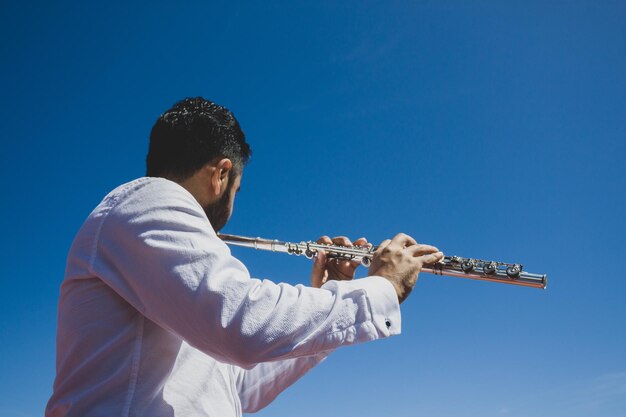 Man playing flute wearing white shirt with sky background