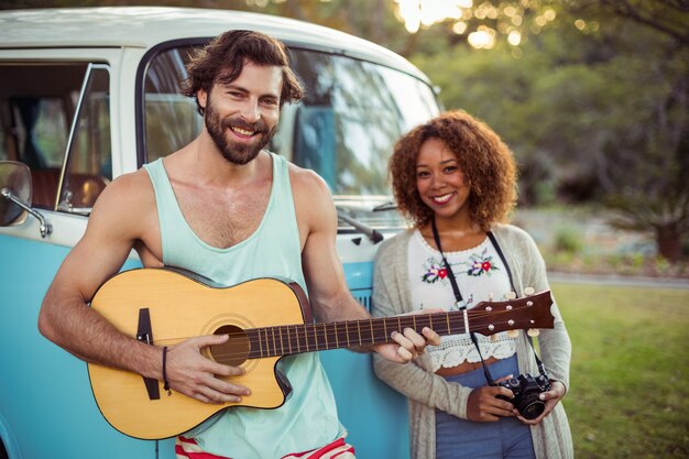 Man playing guitar near campervan while woman standing beside him