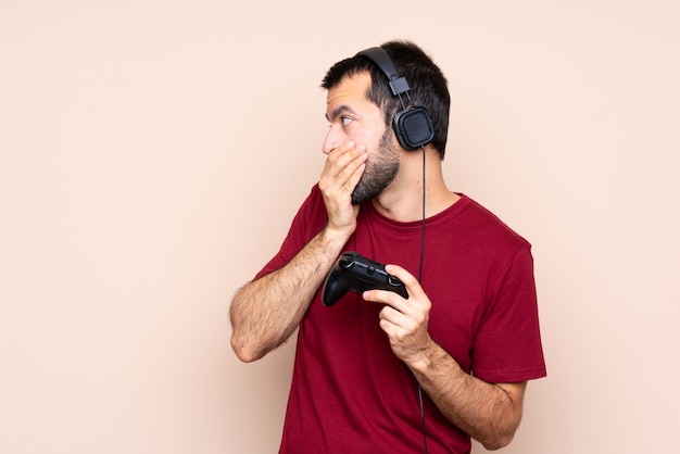 Man playing with a video game controller over isolated wall covering mouth and looking to the side