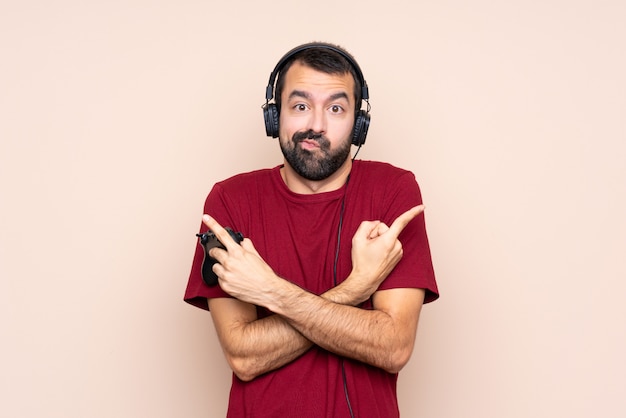 Man playing with a video game controller over isolated wall pointing to the laterals having doubts