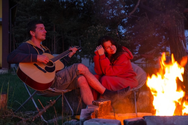 A man plays the guitar a woman listens and sings along A couple in love is sitting by the outdoor campfire in the courtyard of the house on camping chairs a romantic evening