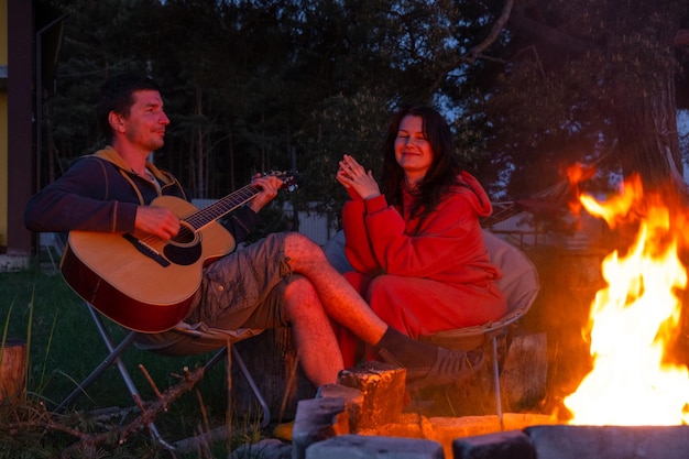 한 남자가 기타를 연주하고, 한 여자가 듣고 함께 노래하고, 사랑에 빠진 한 커플이 야외 캠핑 불 에 앉아, 집 안의 마당에서 캠핑 의자에, 만적인 저녁.