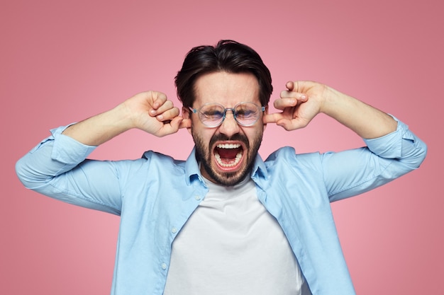 Man plugs ears and screaming opens mouth closes eyes against pink wall