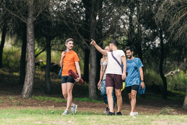 Man pointing ahead walking with friends in the middle of the forest with bags and yoga mats