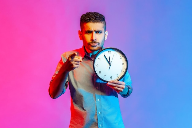 Man pointing finger at camera holding big wall clock in hand time to action serious expression