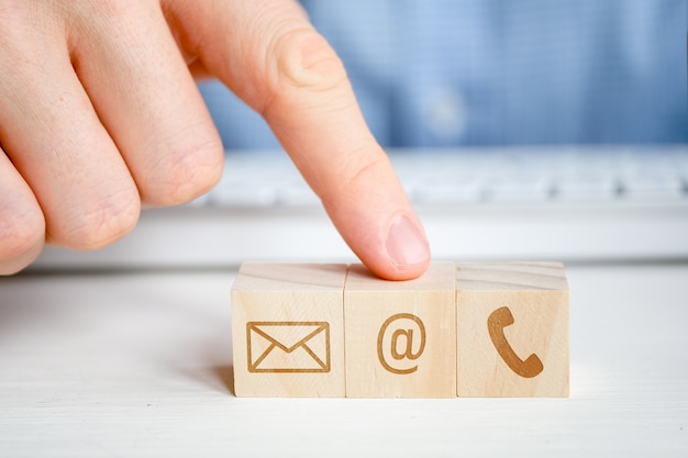 A man points to a wooden cube with the image of an email symbol next to a telephone and a letter with his hand. Contact  for communication.