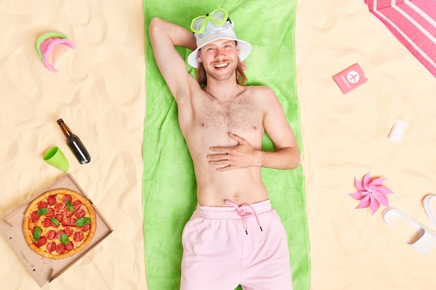  man poses shirtless on green towel wears sunhat and shorts enjoys summer vacation eats tasty snack with beer enjoys summer vacation good rest