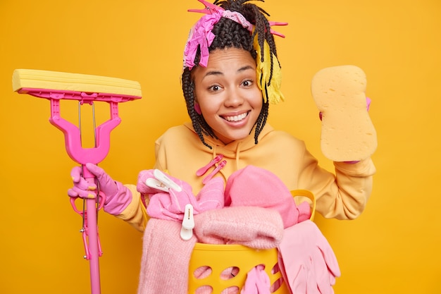 man poses with mop and sponge brings house in order does laundry wears protective rubber gloves isolated on yellow wall